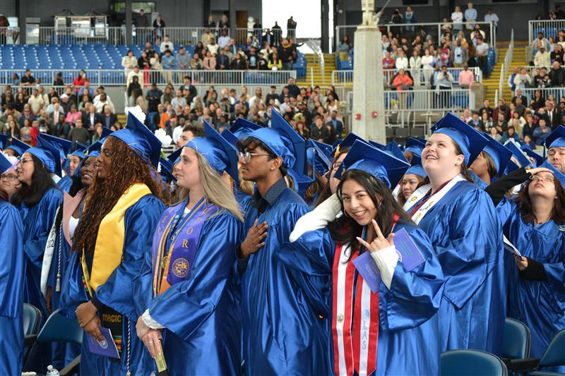 smiling graduate makes hand sign and winks at camera, surrounded by other gradutes
