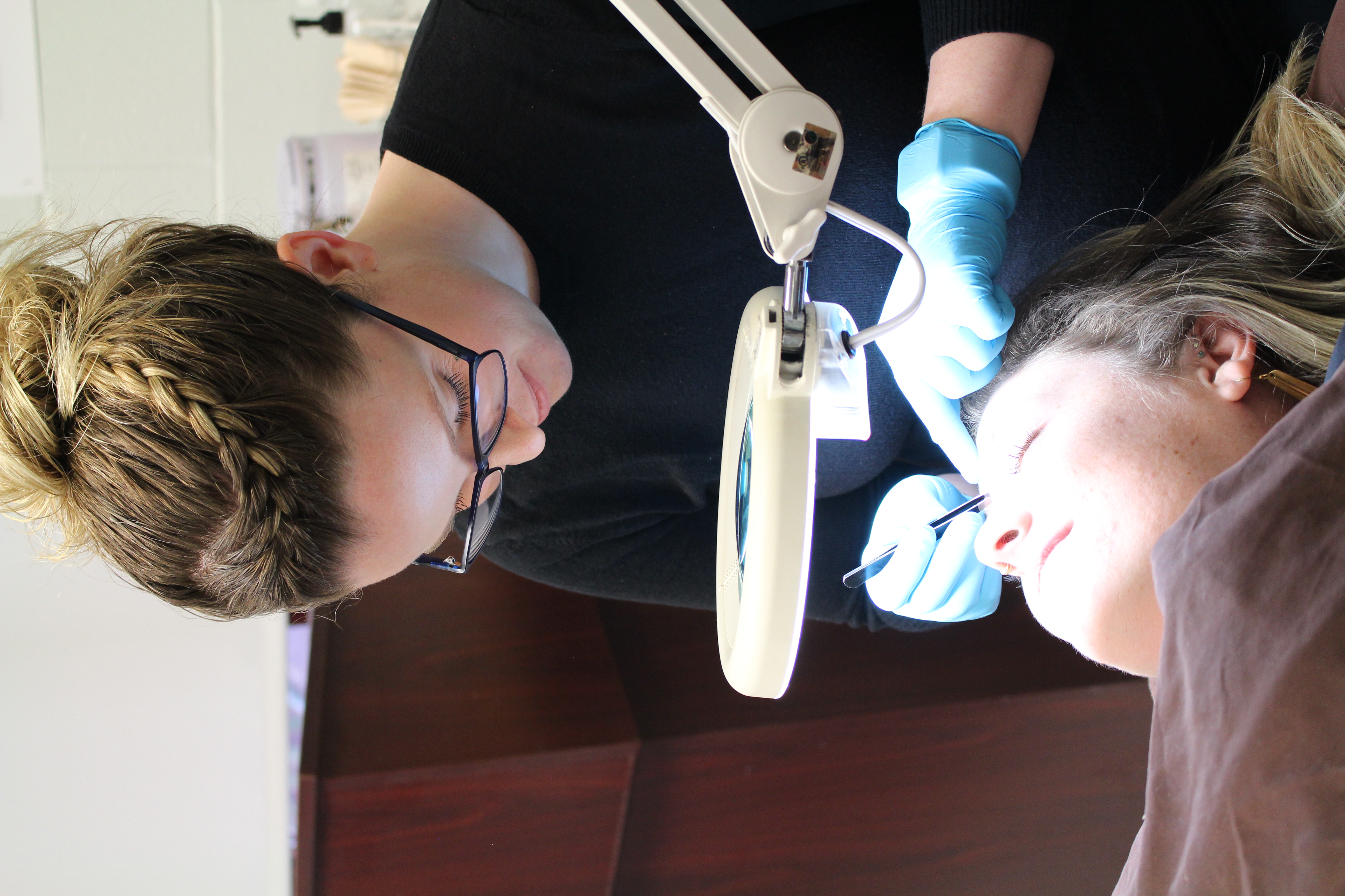 Esthetician student plucks client's eyebrows under lit magnifying glass.