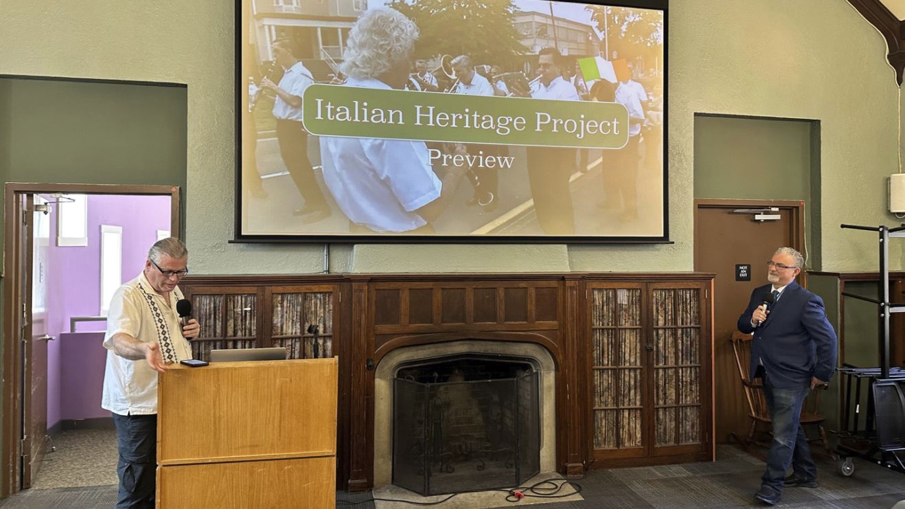 Two men present the "Italian Heritage Project Preview" in a room with a large projecOne man stands at a podium speaking into a microphone, while the other stands to the side holding a mic, near a fireplace.