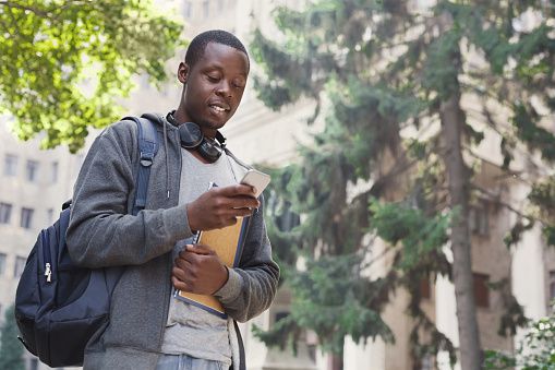 male college student stands outside holding backpack and notebook while looking down and phone with slight smile on face