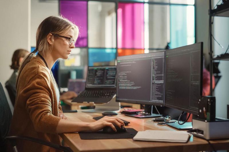 woman concentrates while coding on computer with two screens