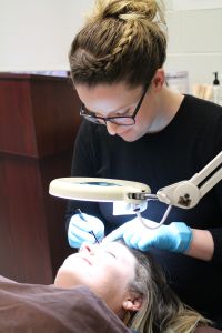 Esthetician student plucks client's eyebrows under lit magnifying glass.