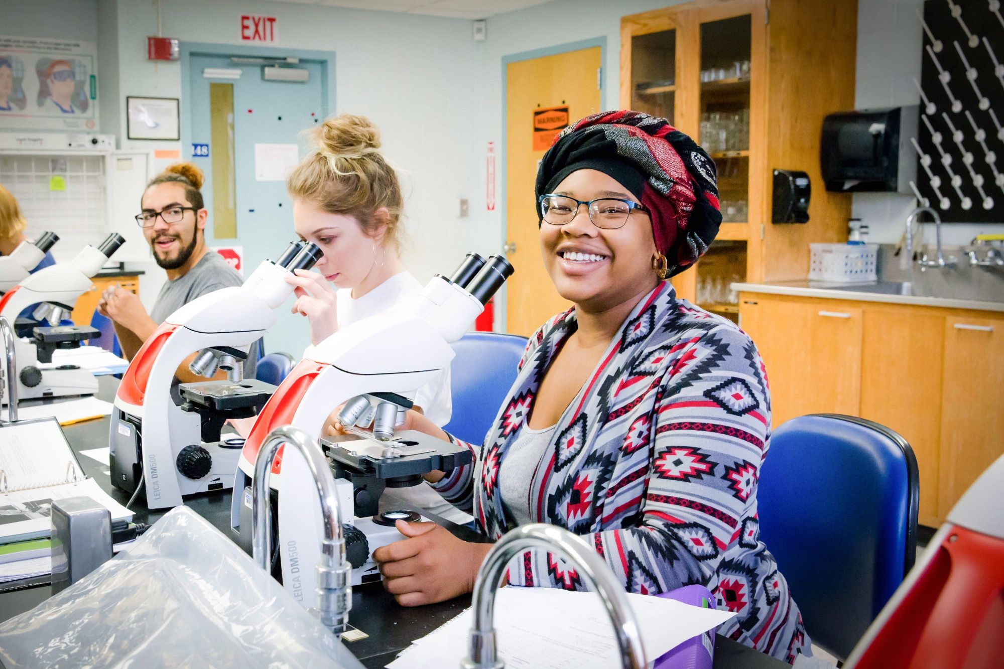 students in classroom use microscopes and look at camera smiling