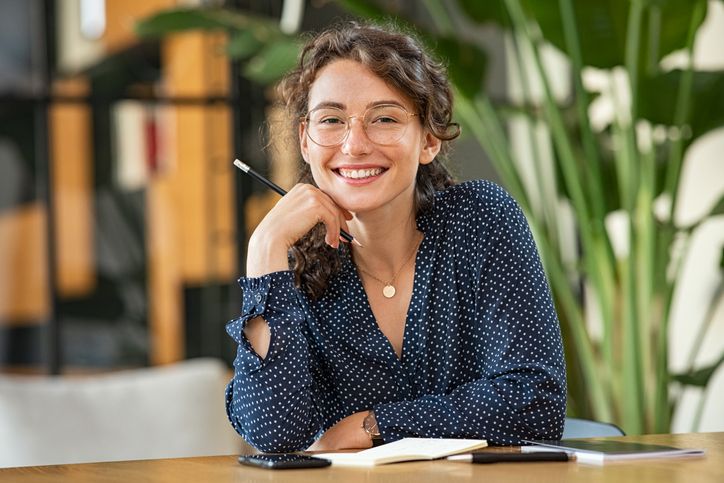 female stuent smiles with hand on chin resting on desk with plants and window behind her.