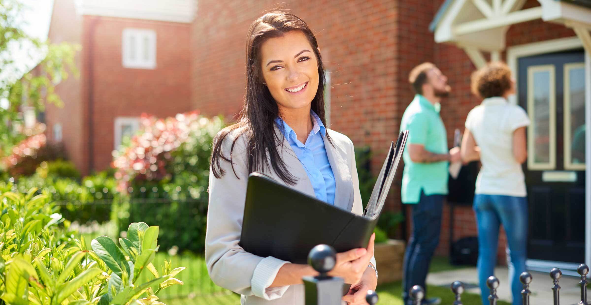 real estate agent holding binder smiles at camera while couple in background looks at house